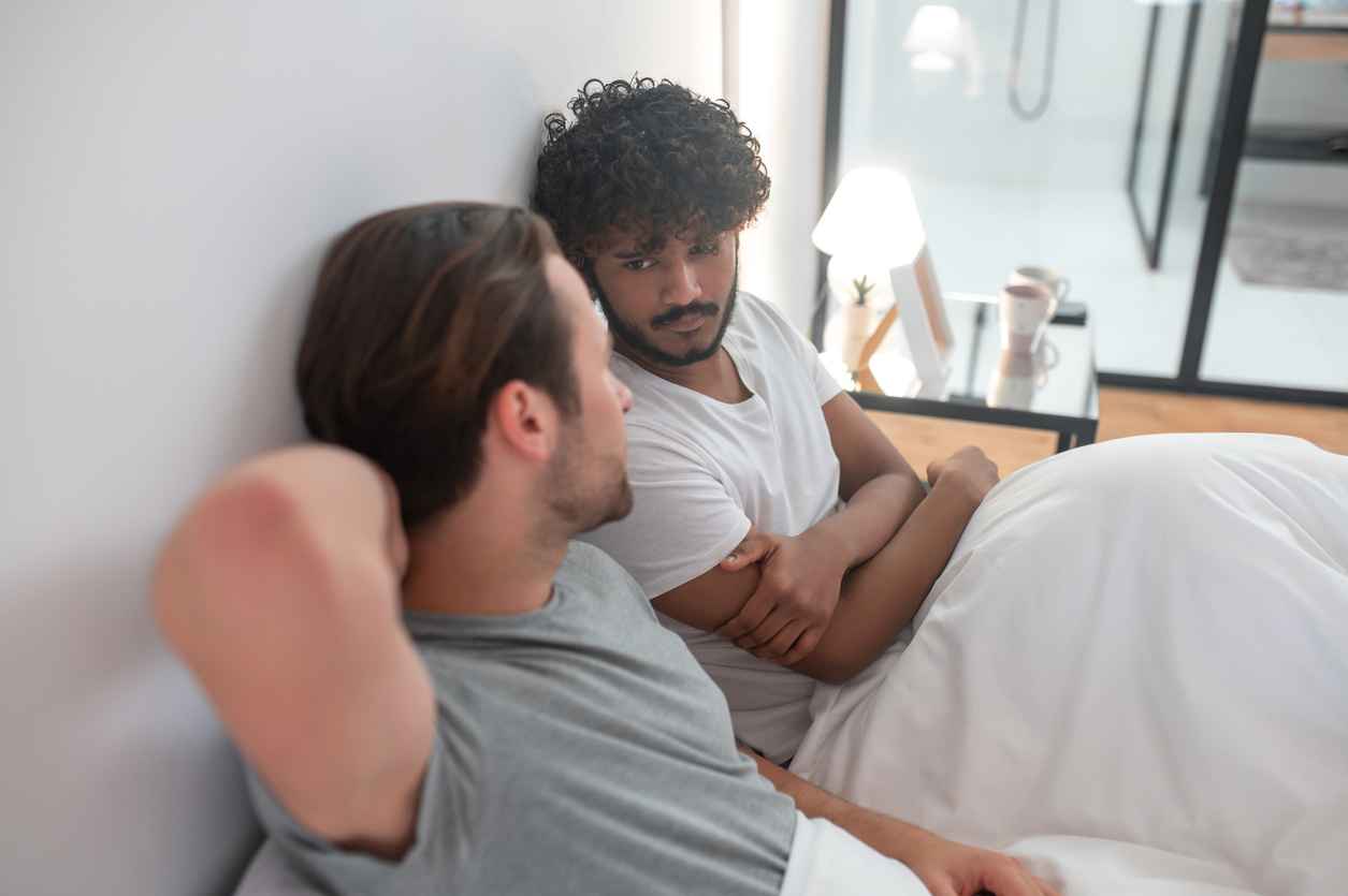 Serious concentrated Indian man seated in bed staring at his Caucasian boyfriend during the talk