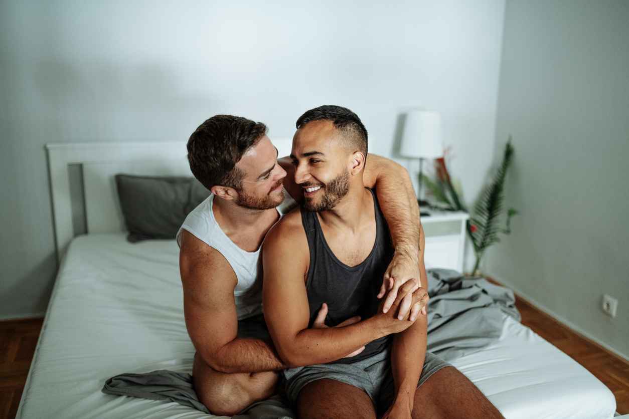 Gay couple smiling and holding hands in bedroom; Tips for Improving Sexual Health