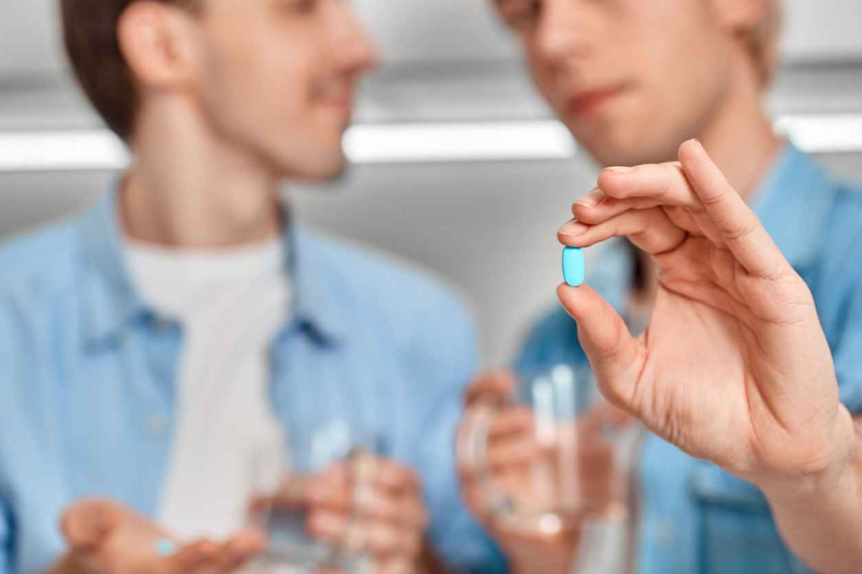 Young gay couple at home standing while holding cups of water and blue pill pre-exposure prophylaxis close-up